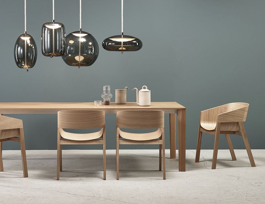 A modern dining setting featuring dark glass pendant lights hanging above a wooden table with matching chairs. The backdrop has a teal wall, and the table is adorned with a minimalist ceramic tea set and glasses.
