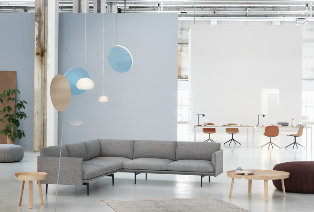 A spacious and modern open-concept workspace with a muted colour palette. On the left, a large light blue wall is adorned with whimsical hanging lights, featuring a combination of soft beige and blue circular panels, some of which have embedded lights. Below is a comfortable L-shaped grey sofa with a small wooden stool beside it. To the right, a transparent table with various office supplies stands, surrounded by a mix of swivel chairs in shades of white and brown. 