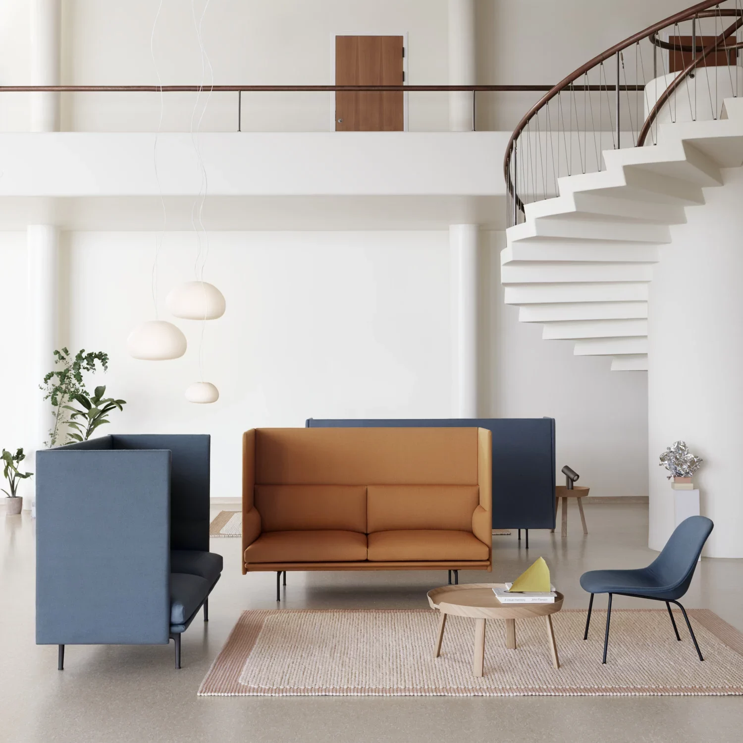 An airy, modern living space featuring a spiral staircase with wooden steps on the right. Below the staircase is a minimalist interior setting with a tan leather sofa and a deep blue armchair, both with contrasting frames.