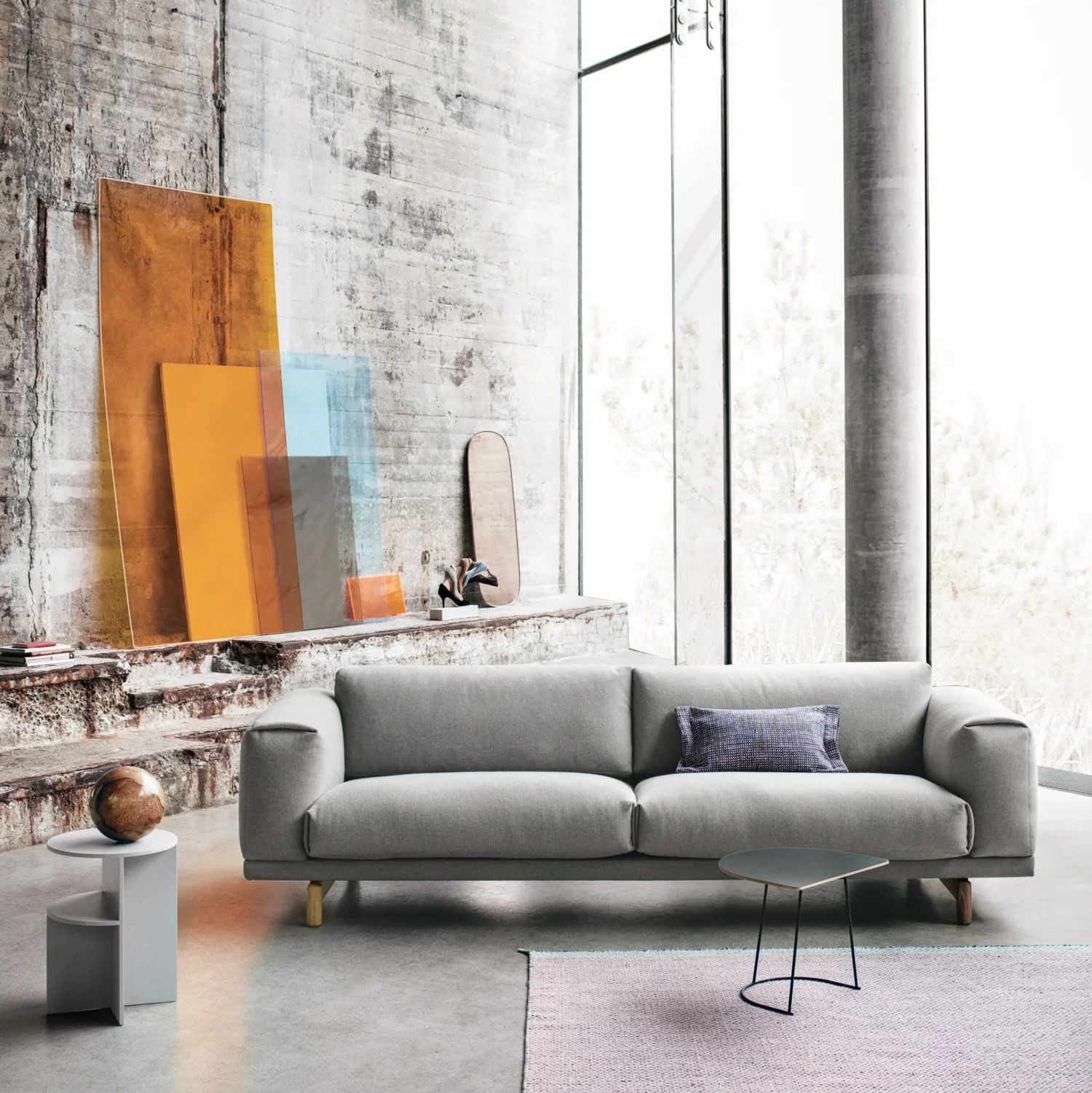 A modern living room with an industrial vibe. The space features a grey sofa adorned with a single purple pillow. In front of the sofa is a minimalistic black metal round table. On the left, against a rustic concrete wall, are large panels of abstract artwork displaying gradients of orange, blue, and brown. A mirror with a rounded top is leaned against the same wall. 