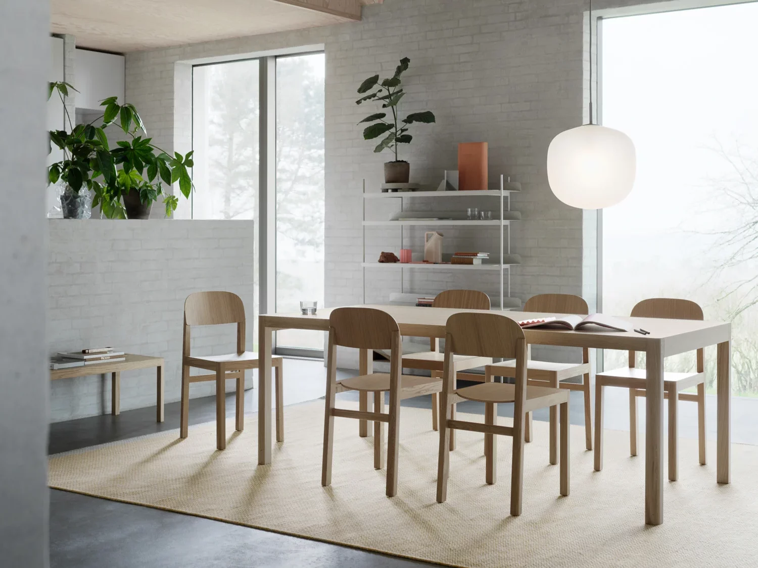 A modern dining area featuring a white brick wall, large windows, and wooden chairs surrounding a rectangular wooden table. A spherical white pendant light hangs above, and an open shelf with decorative items is placed against the wall. A potted plant is positioned near the window.