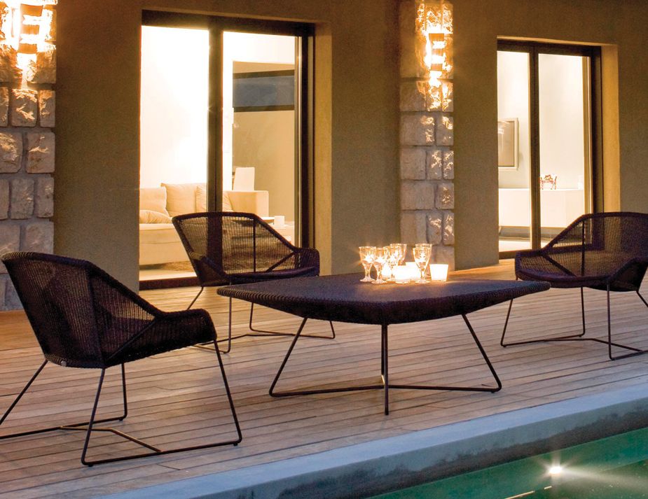 An elegant outdoor seating area with woven, modern chairs and a wooden deck beside a pool. The interior glimpsed through the doorways looks to have a contemporary design, and the setting sun gives the entire scene a warm and inviting ambiance.