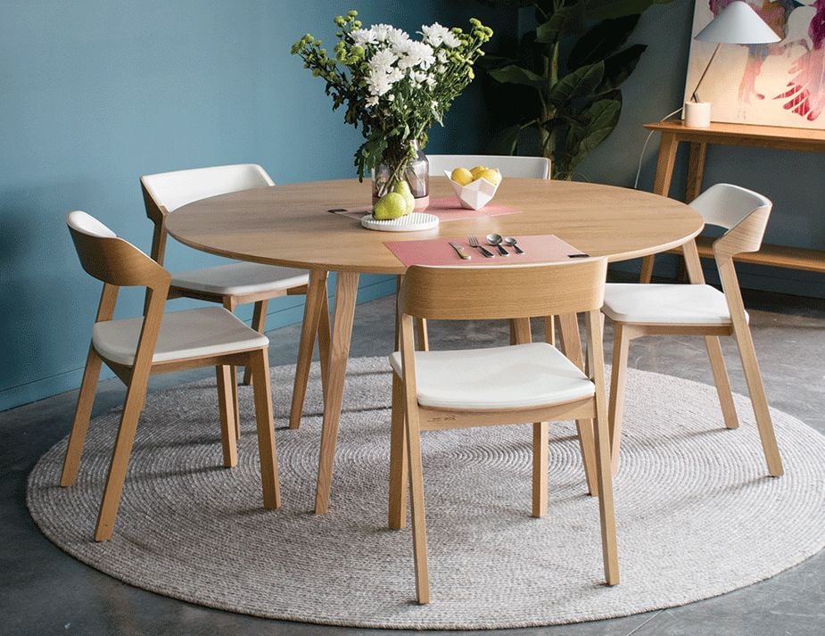 A round wooden dining table set against a teal background. The table is accompanied by four wooden chairs with white cushions. The tabletop has a vase with white flowers, a pink table mat, some fruits, and cutlery. The room features a floor rug and a painting of a pink flamingo on the wall.