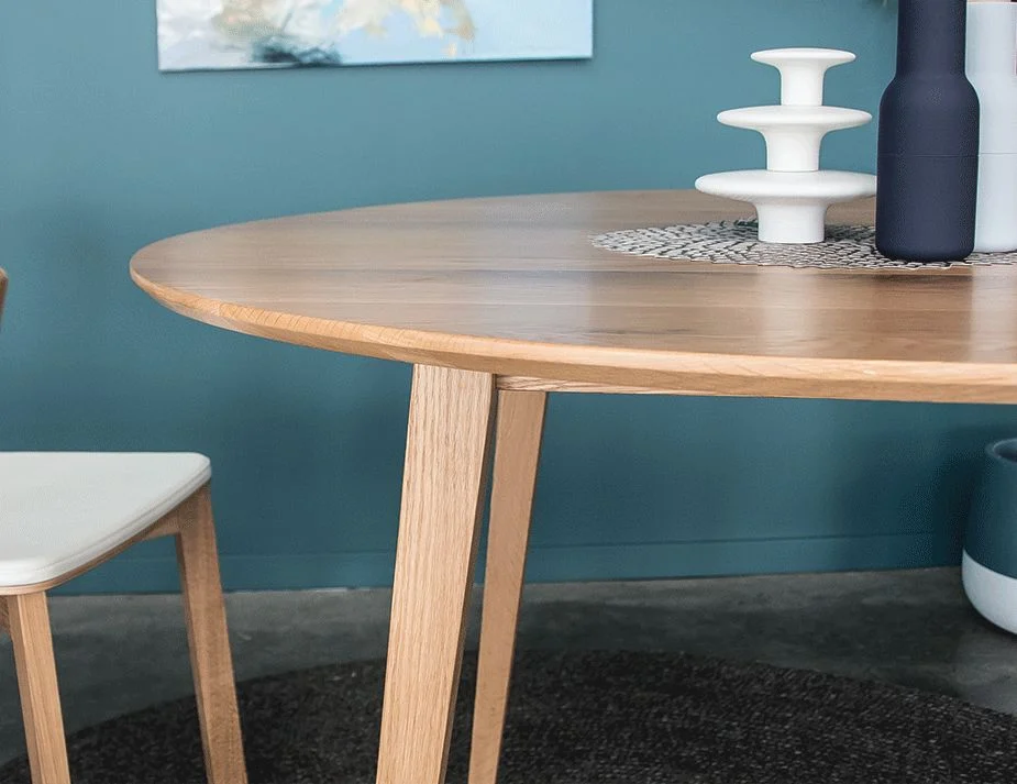 A close-up view of a wooden round table with a natural finish, accompanied by a white chair, set against a teal-colored wall with decorative items.



