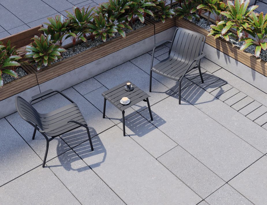 An elevated view of an outdoor terrace with two dark-coloured chairs arranged around a small table. On one side, a long wooden planter box holds an arrangement of green plants with red-tinted leaves. The flooring is made of square grey tiles.