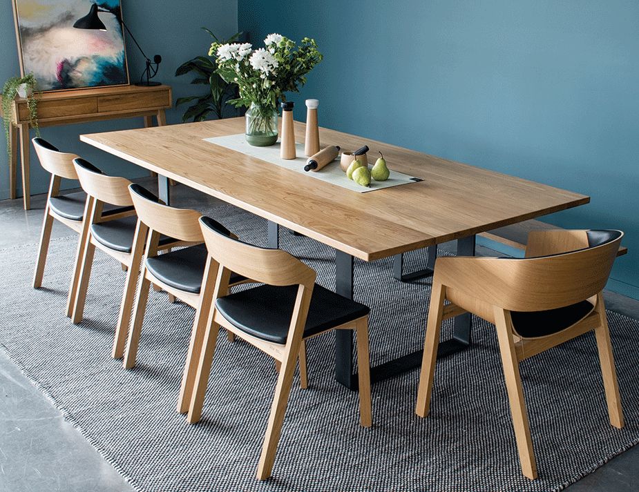 A spacious dining area with a rectangular wooden table set against a blue wall. The table is paired with six wooden chairs that have black cushions. There are various decorative items on the table, including a lamp, a vase with white flowers, and some fruits. A colourful painting and a wooden cabinet are also visible in the room.