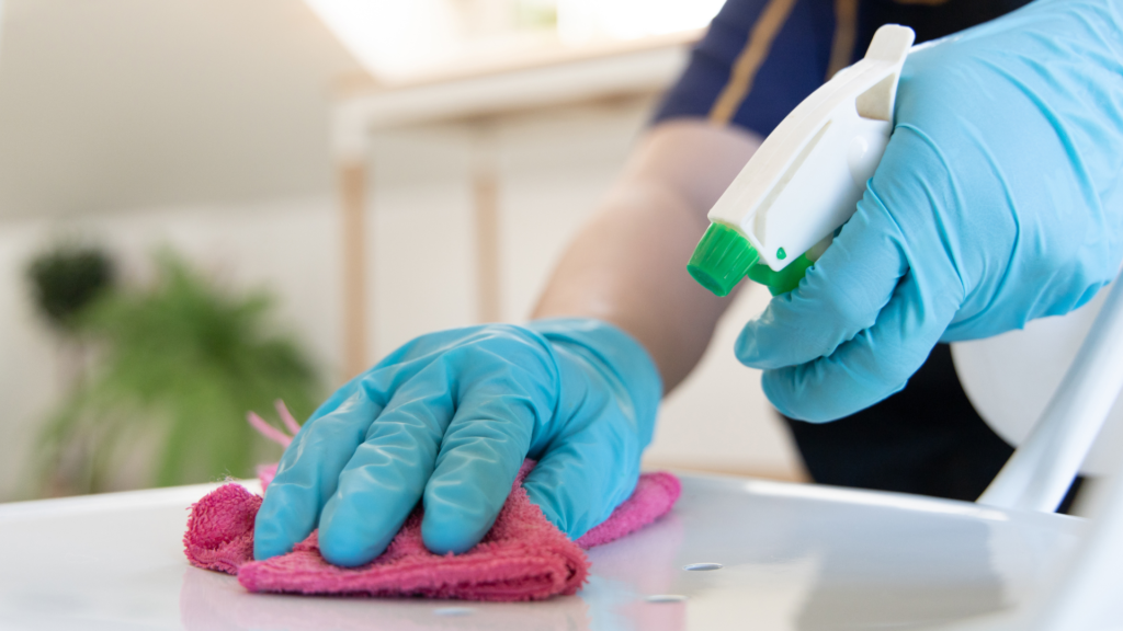 A close-up of a person's hands wearing blue gloves. One hand holds a pink cloth, wiping a white surface, while the other hand holds a cleaning spray bottle with a green nozzle.