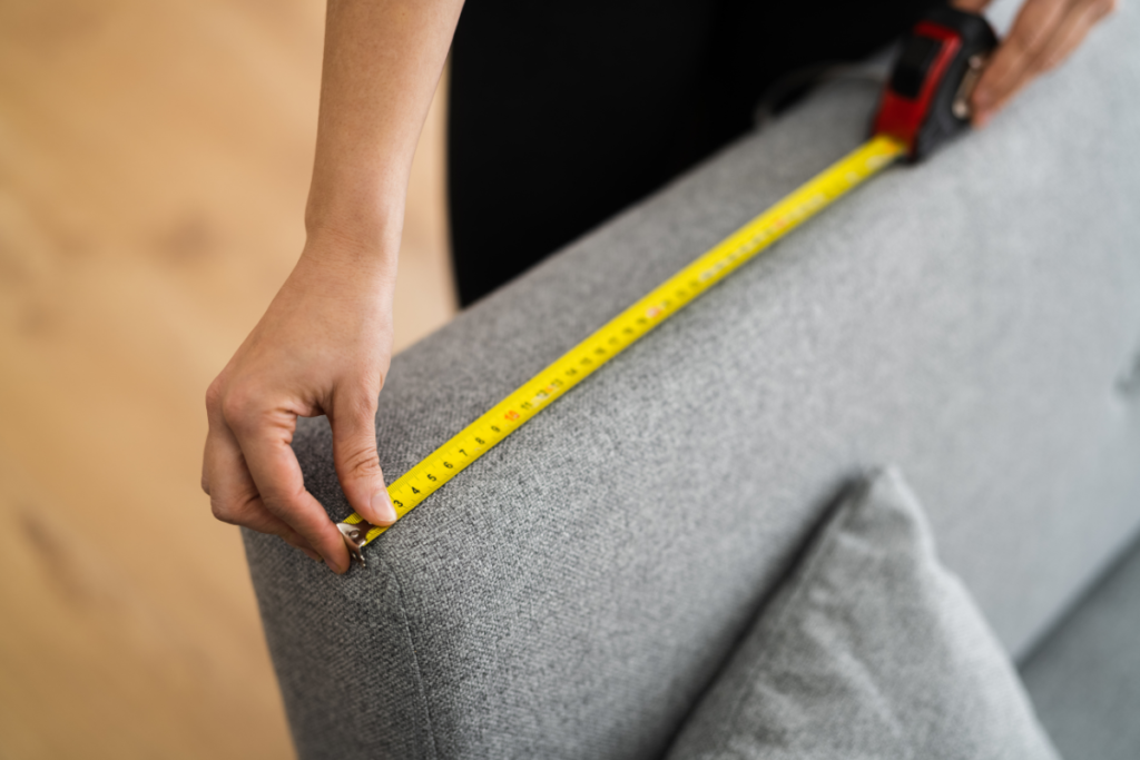 A close-up of a person's hand holding a yellow measuring tape against a grey fabric surface, possibly a piece of furniture, illustrating "how to choose a sofa." The measuring tape is extended and is being used to measure the width of the fabric.
