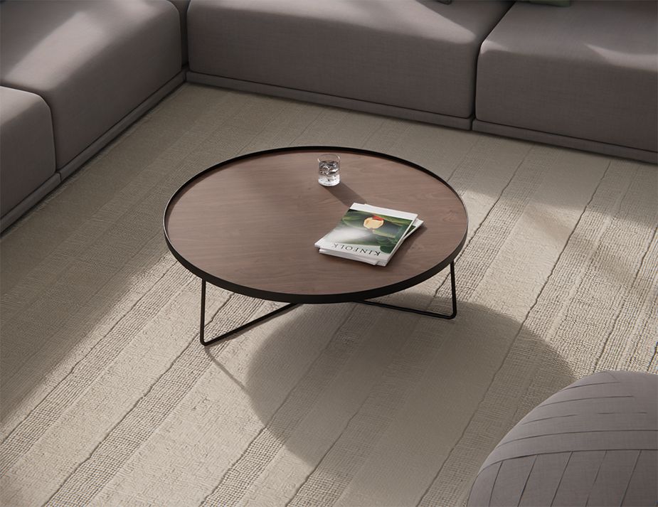 A minimalist living room with a round wooden coffee table placed on a beige textured rug, flanked by a grey sectional sofa. 
