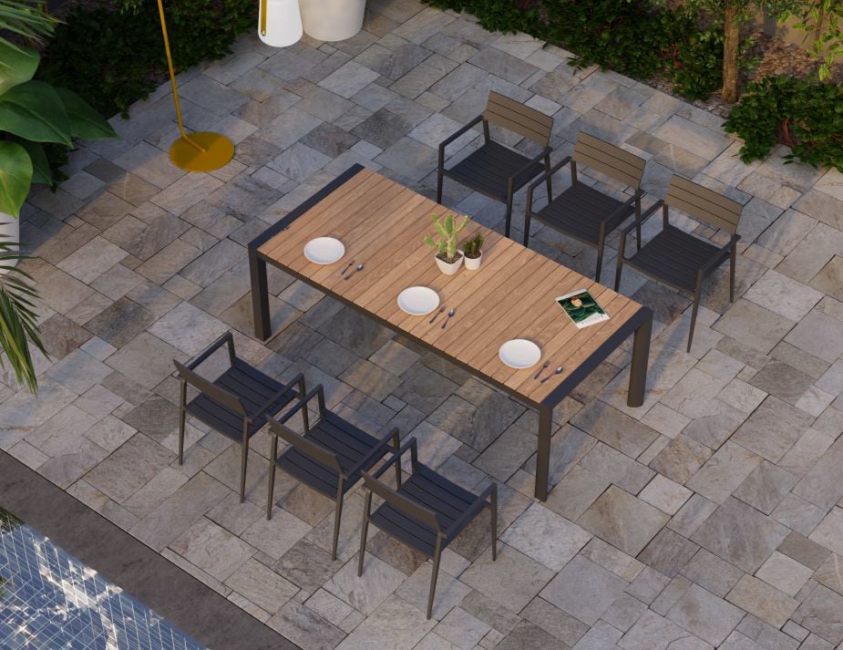 An outdoor dining area set on multicolored stone tiles. A wooden table with place settings for four stands in the centre, surrounded by four dark-coloured chairs. A yellow floor lamp stands to the left, and the background showcases lush green plants and a partial view of a pool.