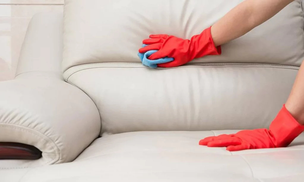 A person wearing red gloves is cleaning a white leather sofa with a blue cloth.
