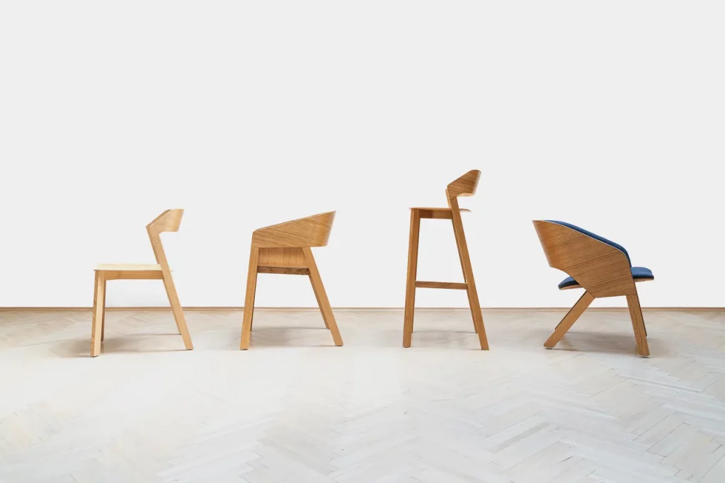 A minimalist room featuring three uniquely designed wooden chairs. From the left, the first chair is a sleek low-backed design with a cut-out silhouette. The middle chair is a taller stool-like design with an angular backrest. The third chair on the right combines the features of the first two with a curvilinear backrest and a cushioned blue seat. All chairs are set against a pristine white backdrop with a light wooden herringbone floor.
