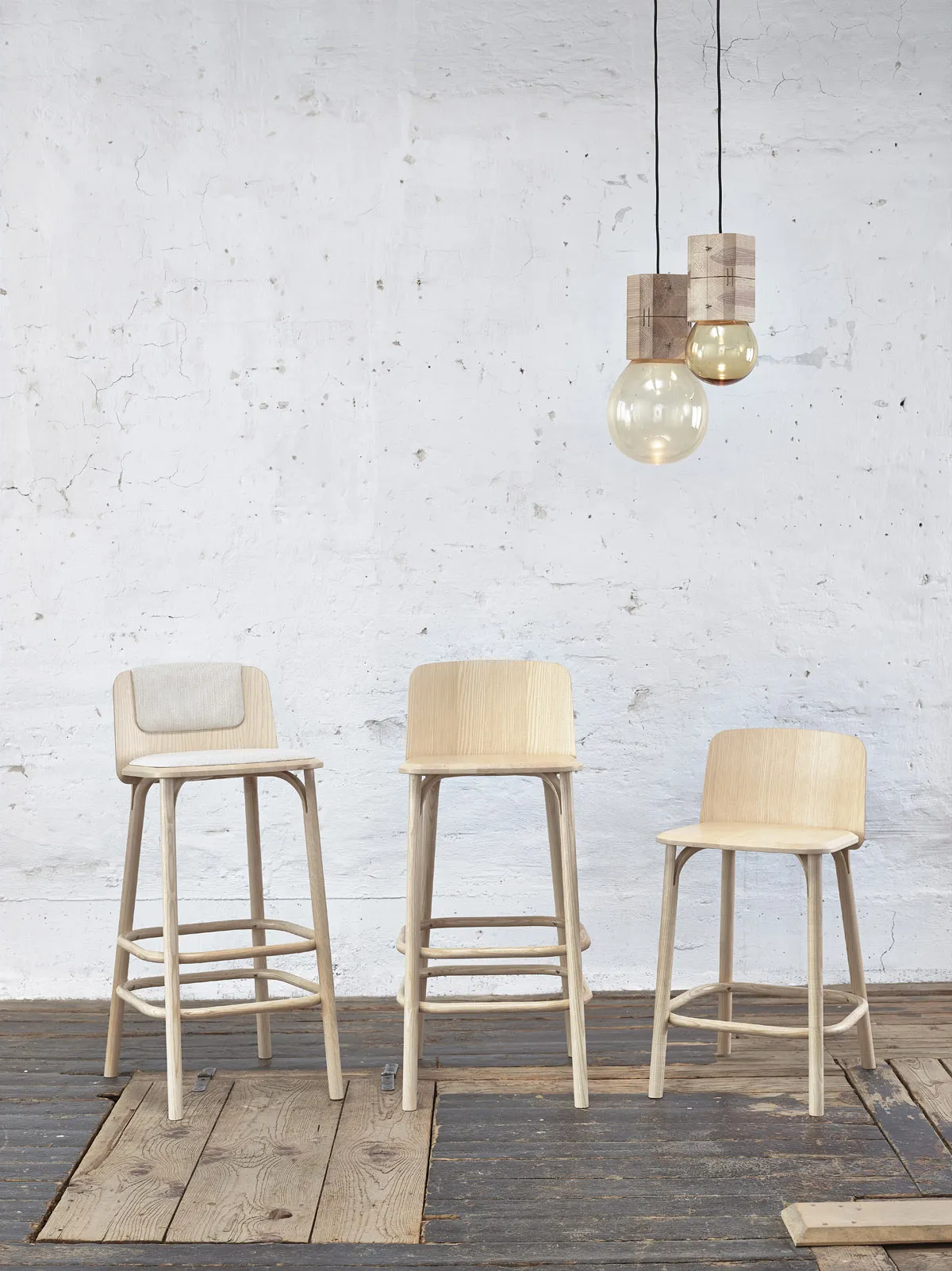 Three wooden bar stools of varying heights positioned on rustic wooden flooring against a textured white wall, accentuated by two hanging pendant lights with wooden fixtures and clear glass bulbs.