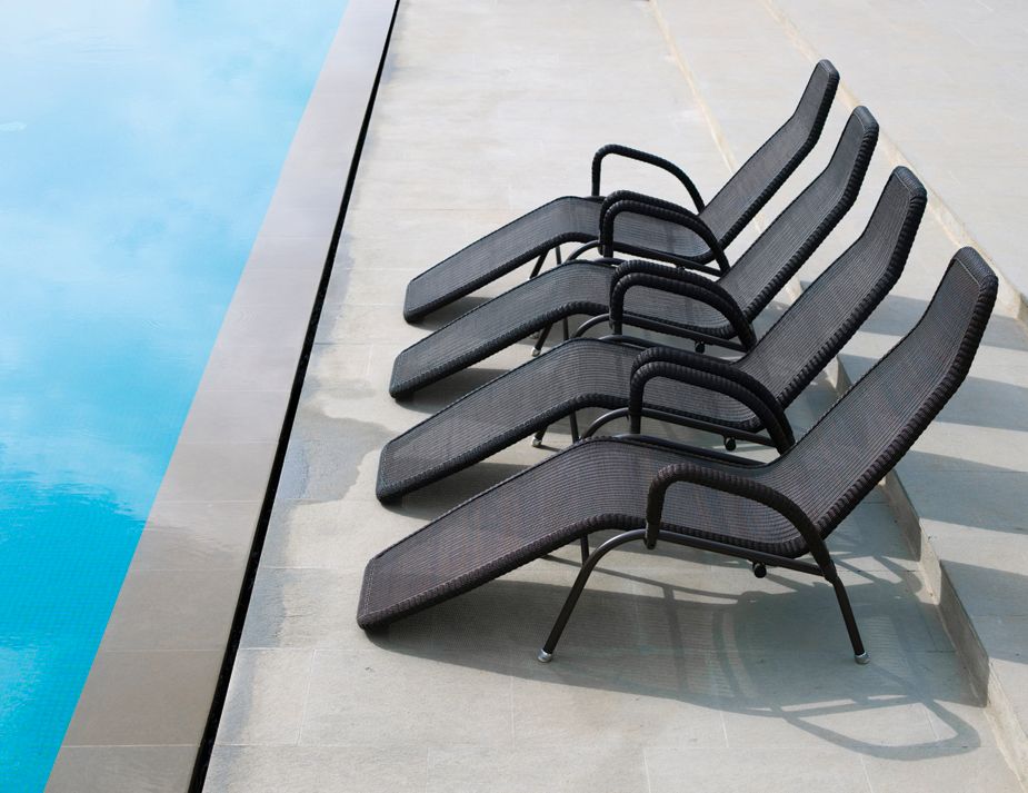 A row of sleek, black woven lounge chairs arranged beside a tranquil swimming pool with crystal-clear blue water. The chairs, featuring curved armrests and ergonomic design, sit on a smooth light-coloured stone deck, casting long afternoon shadows.