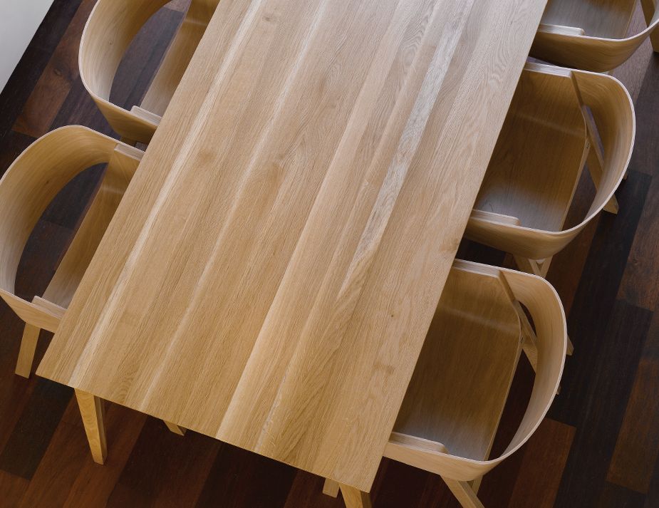 A top-down view of a wooden dining table with matching curved chairs. The surface showcases the beautiful wood grain, and the floor has a contrasting darker wooden finish.



