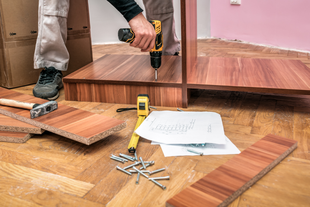 A close-up of a person in the process of assembling a wooden shelf. The person holds a power drill, drilling into the wood. Surrounding the workspace are various tools such as a hammer, measuring tape, and screws. Wooden planks and a blueprint of the shelf lie scattered on the wooden floor. Only the hands and legs of the person are visible, emphasizing the assembly process.




