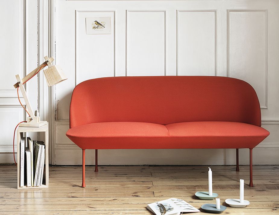A minimalist living room setting featuring a vibrant red, curved sofa with slender legs, placed against a white paneled wall adorned with a framed bird illustration. To the left of the sofa is a wooden adjustable desk lamp with red cords, resting atop a small white table that holds a few vertically stored magazines.