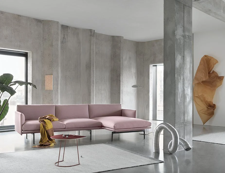 A modern living room showcasing a pink sofa against a raw concrete wall with a floor lamp beside it. An abstract metallic sculpture is placed on the floor, and a large draped brown paper decorates another section of the wall.

