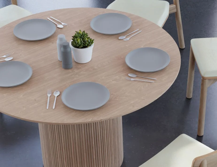 A close-up view of a round wooden dining table with light grey plates, silverware, and a white potted succulent plant. The base of the table has a ribbed design, and off-white cushioned chairs surround it.

