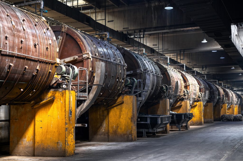 Large cylindrical industrial machinery, possibly rotary dryers or kilns, aligned in a row inside a dimly lit factory setting. The machines have a rusted brown appearance and are supported by yellow concrete bases.


