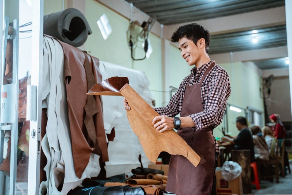 A young man in a workshop, wearing an apron and holding a piece of leather, examining its quality with rolls of leather material stored in the background.
