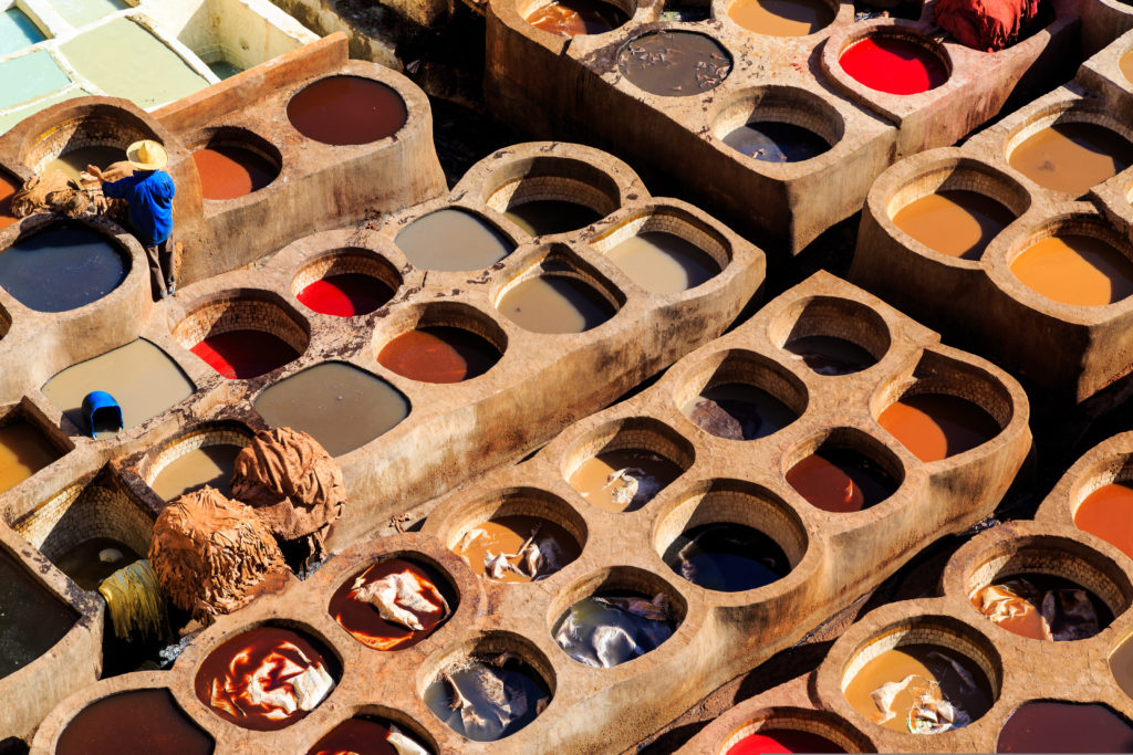 An aerial view of a traditional tannery with multiple circular stone vats filled with various coloured dyes and liquids. A worker in blue attire and a hat is seen tending to the hides, which are being dyed in the vats. The tannery displays a vibrant array of colours from the different dyeing processes.