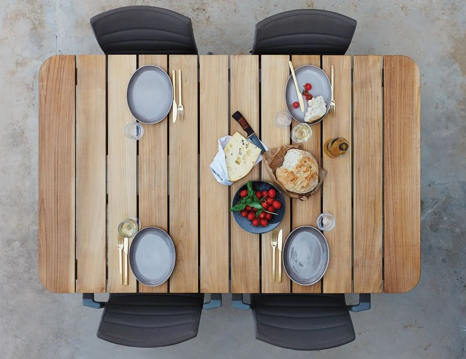 An overhead view of a rectangular wooden dining table set outdoors. The table is laid with plates, glasses, cutlery, a bottle of wine, a basket of bread, and dishes with cheese and strawberries.


