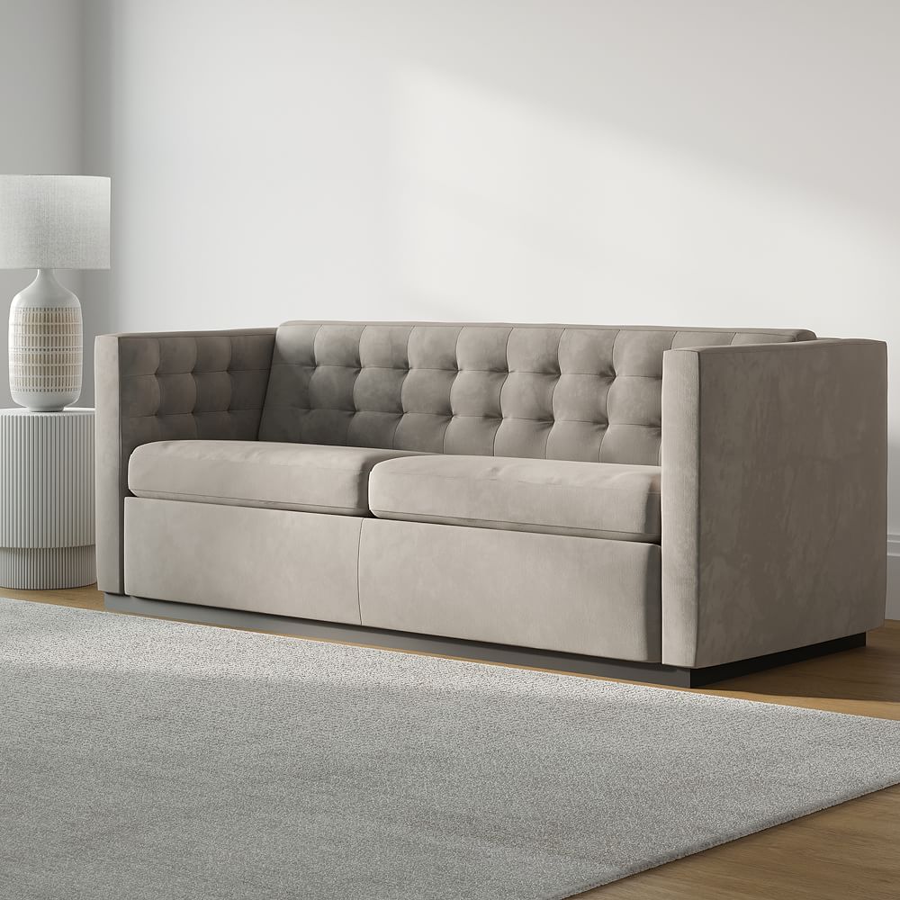 A minimalist living room design with a light gray tufted sofa and a cylindrical white table lamp placed on a wooden pedestal. The room has a soft-toned rug and a bright ambiance.

