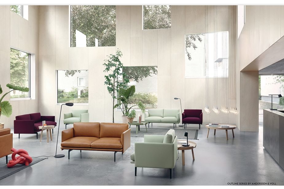 A spacious, modern lounge area illuminated by natural light pouring in from tall, geometric windows. The room boasts an array of mid-century modern furniture, including a tan leather couch in the forefront, mint green armchairs, and a deep burgundy chair off to the left.