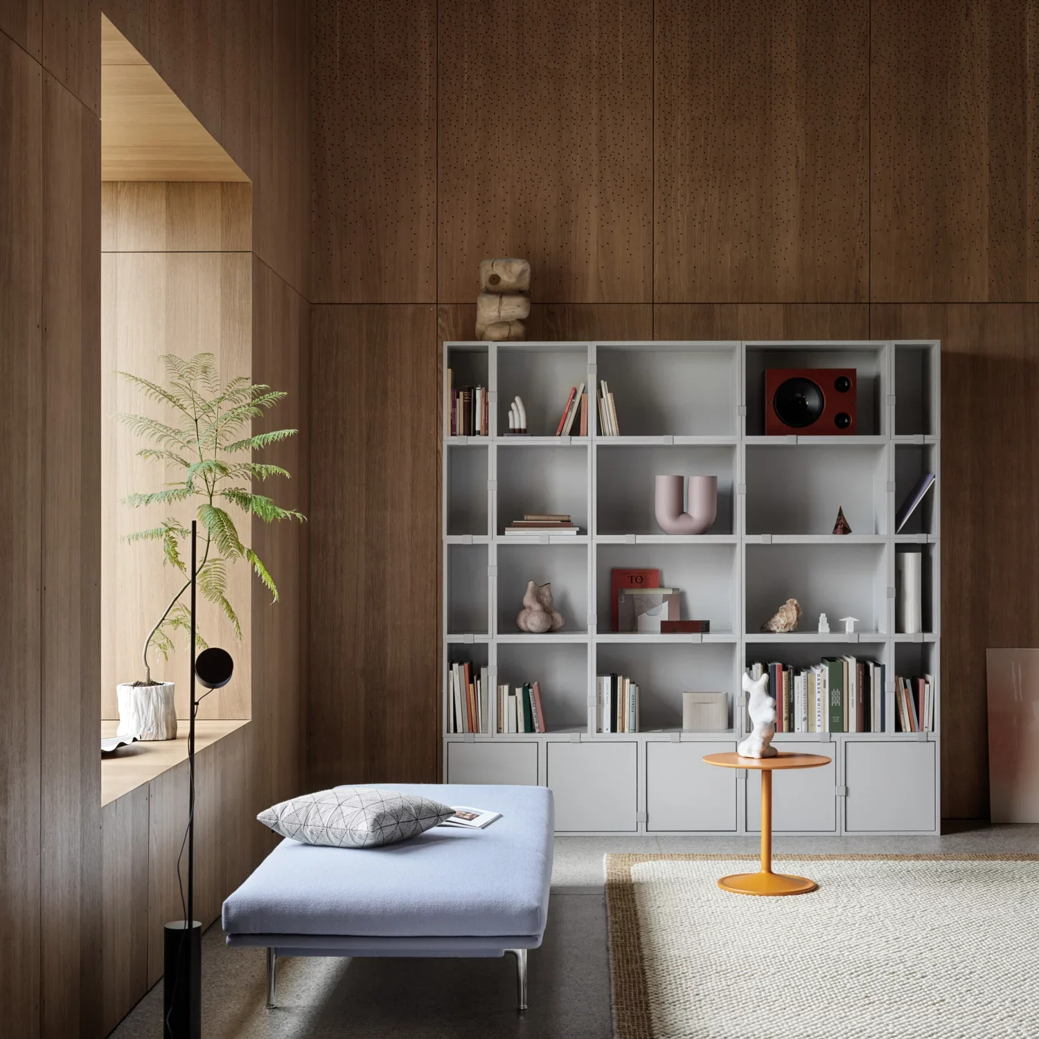 A sophisticated wooden bookshelf unit filled with assorted books and decor items against a wooden-paneled wall. In front lies a muted blue daybed with a cushion, next to a small orange side table.