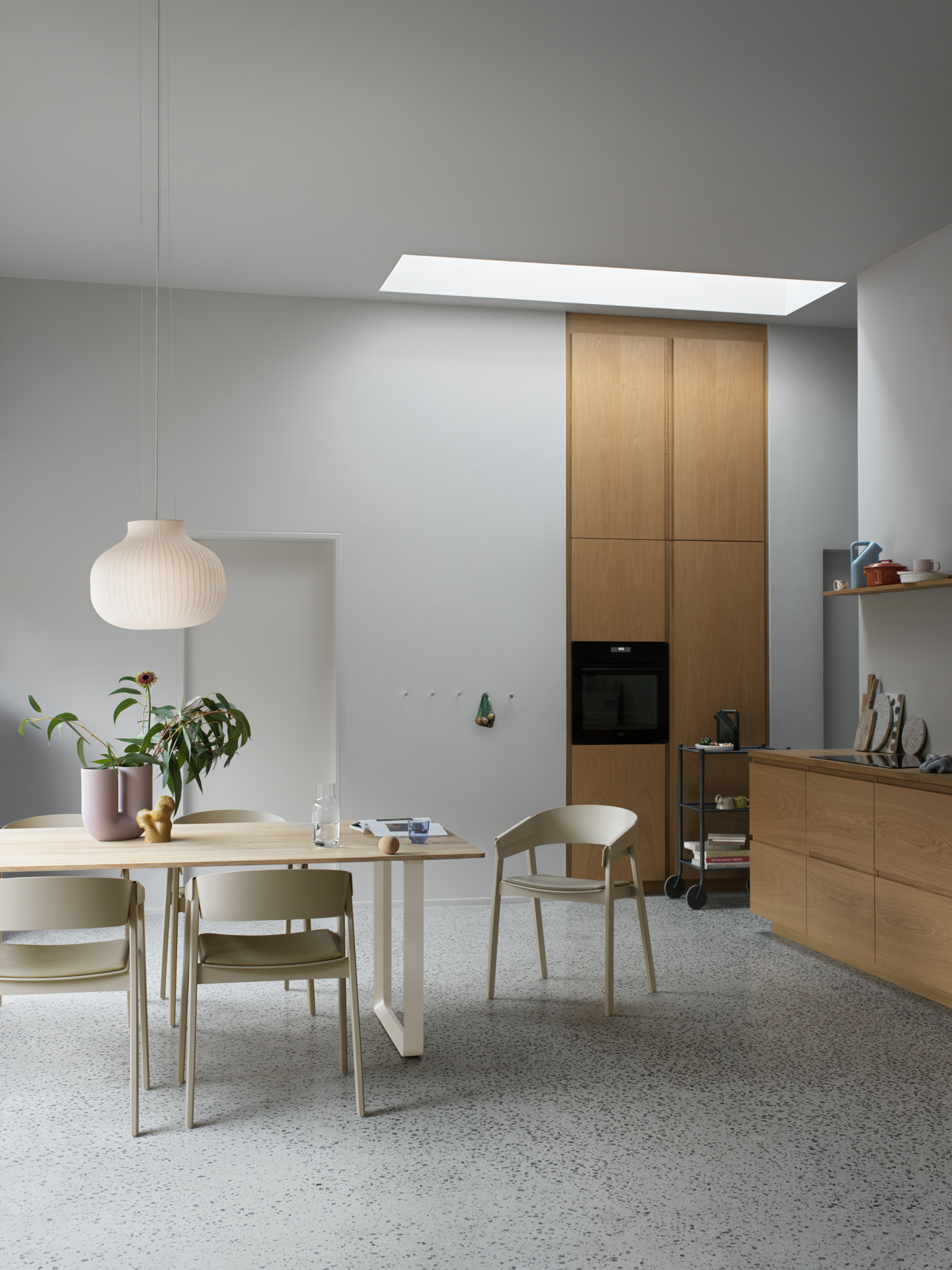 A bright and minimalistic kitchen and dining space with neutral tones. Features include a white pendant lamp, light wood dining table and chairs, and terrazzo flooring. The backdrop consists of a modern kitchen setup and a wooden cabinet