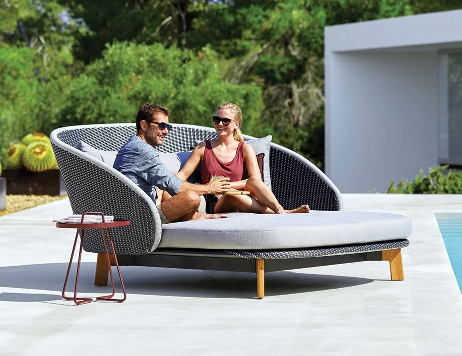 Two individuals, one male and one female, lounge comfortably on a modern, crescent-shaped outdoor furniture near a pool. Both are wearing sunglasses and sharing a conversation, with the woman holding a drink in her hand. A small side table with a book is next to them. 