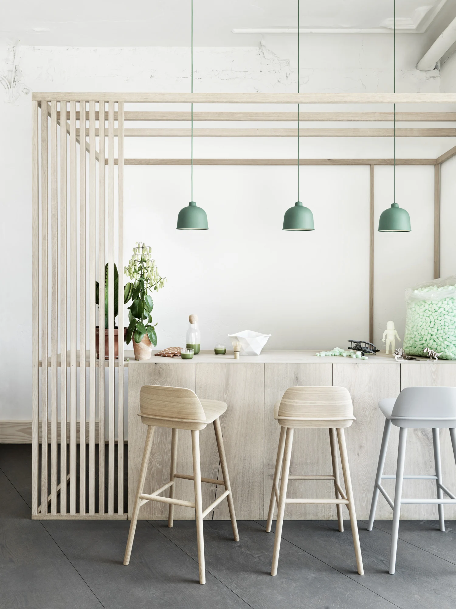 A minimalist kitchen interior featuring a wooden partition with vertical slats, complemented by a light wooden countertop. Above the counter hang three pastel green pendant lights in a row.