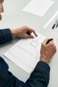 A designer sketching a detailed drawing of a pendant light on paper, with pens and other drafting tools nearby on a gray table.