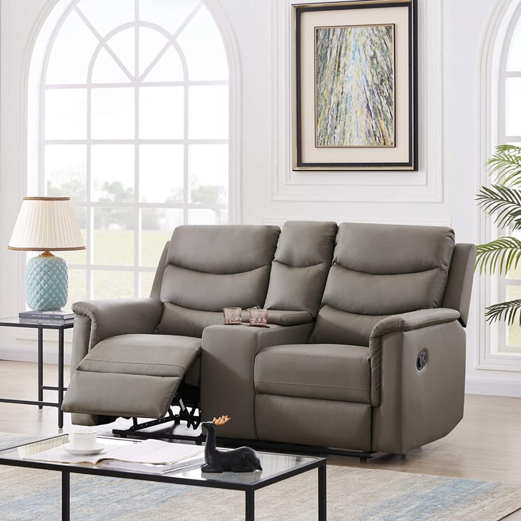 A classic room setting featuring a beige reclining sofa with an extended footrest, a glass coffee table, a lamp, and an artwork on the wall.
