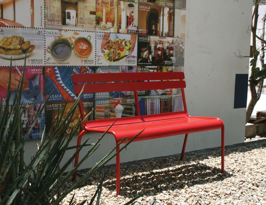 A vibrant red bench sits in an outdoor setting, positioned in front of a wall adorned with colourful stamps depicting various aspects of Oaxacan and Mexican culture, from architecture to food. To the left, sharp green foliage contrasts against the white gravel and stamps, while the right side showcases a slender tree against a white background.