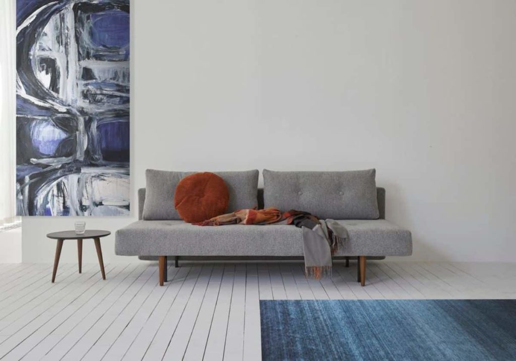A minimalist styled room with a light-colored modern sofa, an abstract wall painting, a small side table, and a textured blue rug.
