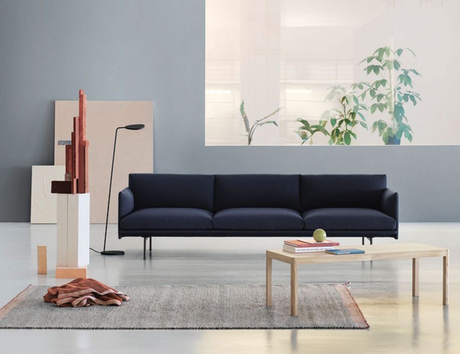 A modern living room setting with a navy blue couch, wooden coffee table, artistic wooden sculpture, and a standing lamp, with a view of plants by the window in the background.



