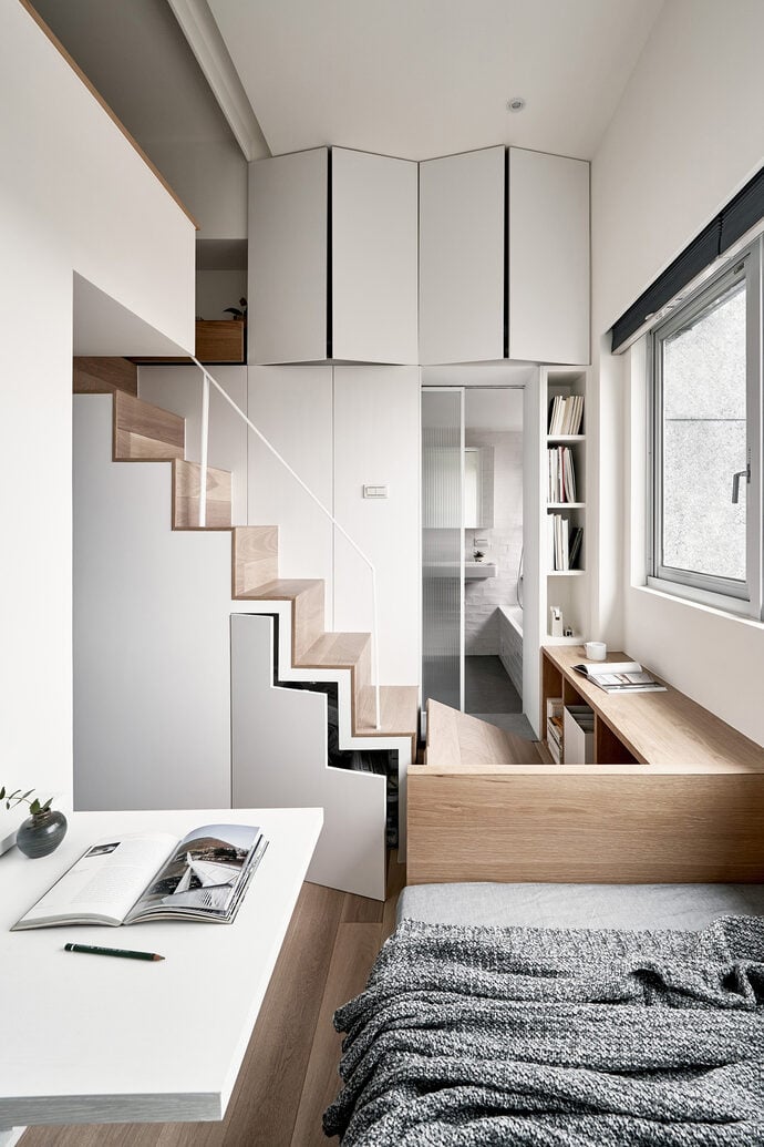 This is an image of a modern interior space. The room features a geometric staircase with wooden steps. Above the stairs, there are white storage cabinets. On the right, there is a desk area with shelving, a window, and a white table with an open book and a pen on it. In the foreground, there's a portion of a bed with a grey knitted blanket.