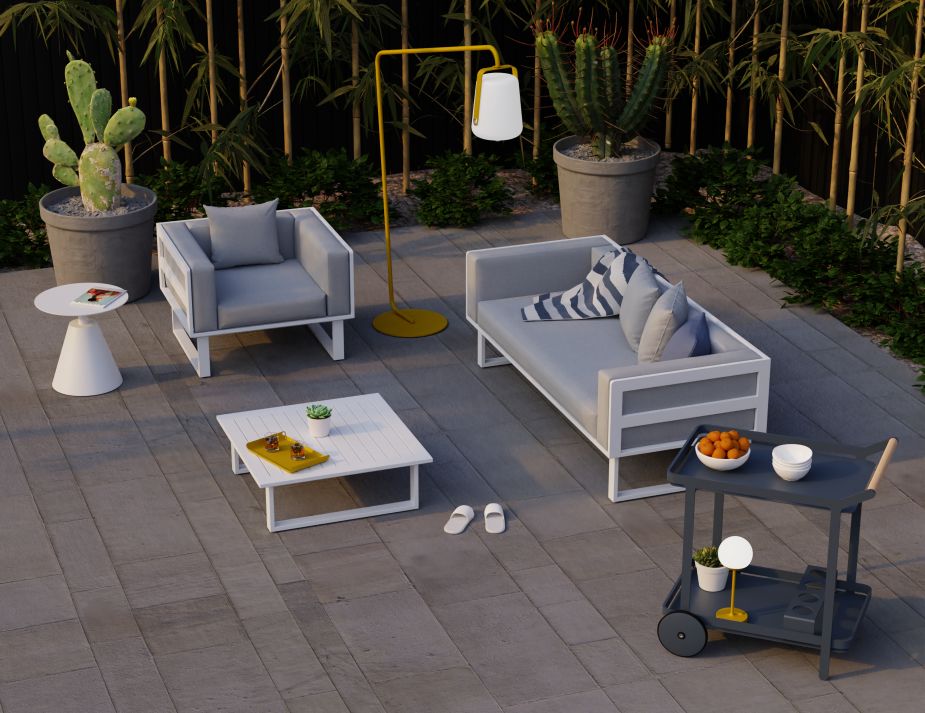 A sophisticated outdoor lounge area during twilight. Two comfortable light gray furniture pieces, a chair and a sofa, are arranged on a patio. A white modern coffee table stands between them, adorned with a book, a small plant, and a tray with a drink. To the side, a movable dark gray cart holds cups and a white lamp. 