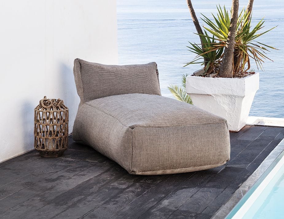 A tranquil oceanfront setting featuring a plush gray chaise lounge placed on dark wooden flooring next to a white wall. Beside the chaise is a rustic woven lantern, while a potted spiky plant sits atop a white ledge overlooking the serene blue ocean.