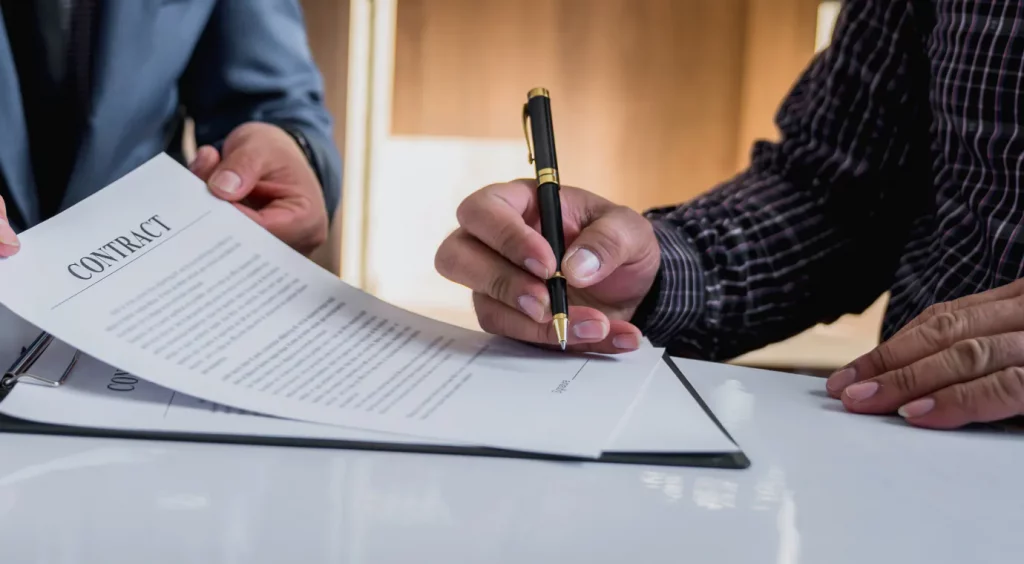 Two individuals at a table, one handing over a contract to the other. 