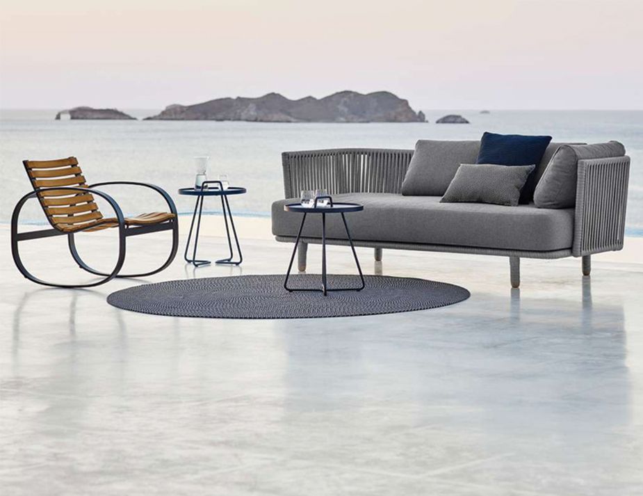 A tranquil seaside setting with contemporary outdoor furniture. A rocking chair with wooden slats and a unique circular frame stands to the left. In the center, a gray upholstered sofa adorned with plush cushions rests atop a textured round rug. Nearby, two sleek metal side tables, one holding a book, complement the modern aesthetic. The calm sea and distant rock formations serve as a serene backdrop.