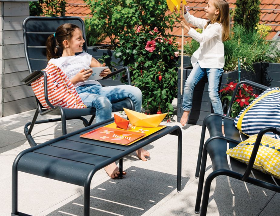 A sunlit outdoor furniture setting featuring two young girls enjoying themselves on a patio. One girl sits comfortably on a modern black chair with an orange-striped cushion, engrossed in a tablet. Beside her, a matching black table displays colourful paper crafts. The other girl stands joyfully, releasing a yellow paper boat into the air.